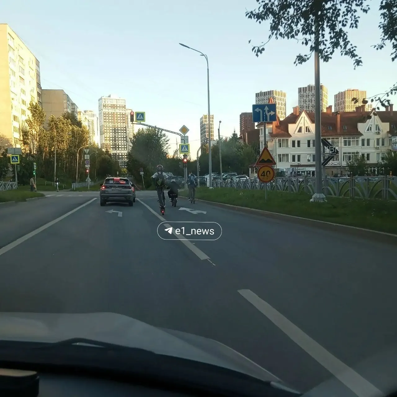 Подростки на самокатах двигаются по дороге на перекрёстке, вызывая риск аварий и тревогу у прохожих.