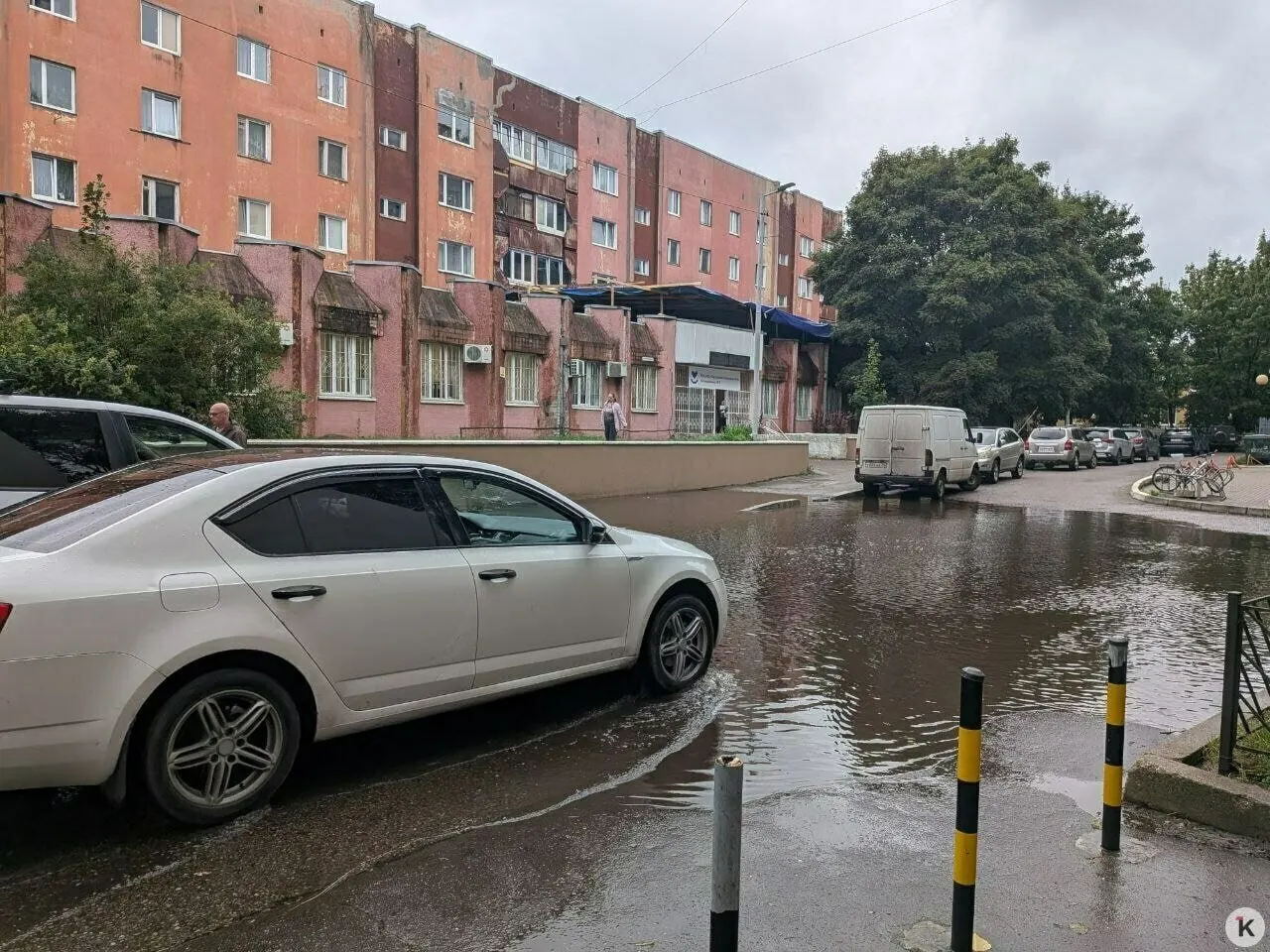 Дождь затопил подход к поликлинике: лужа перед входом настолько велика, что пройти невозможно, люди обходят её по газону и через забор.